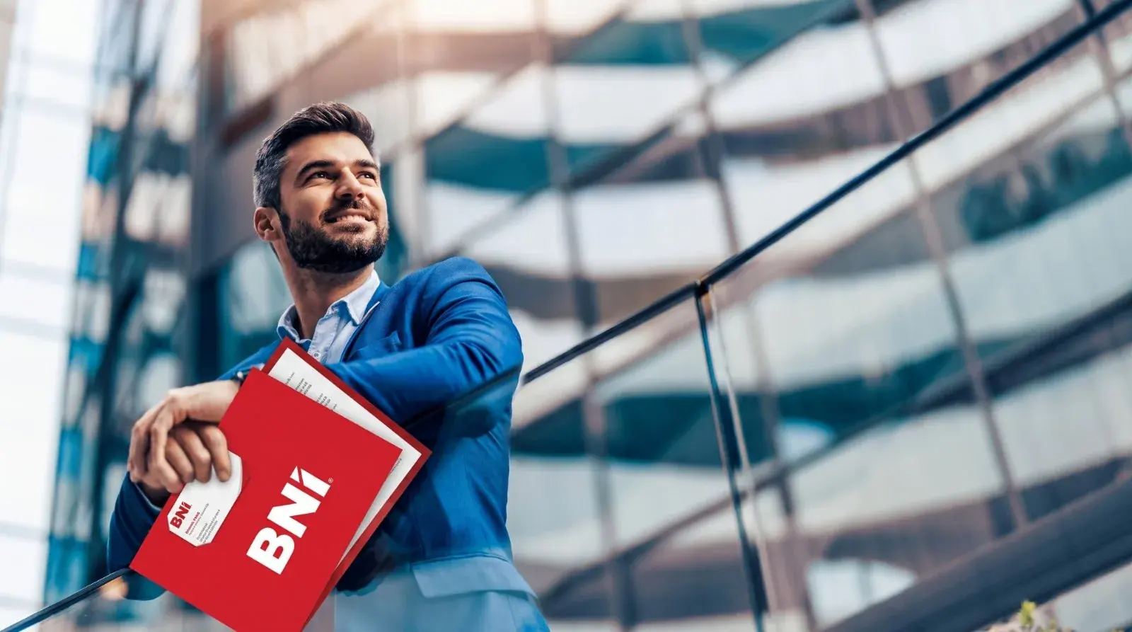 Mężczyzna w garniturze z czerwoną teczką BNI przed przeszklonym biurowcem