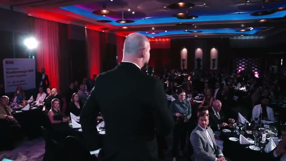 Konferencja BNI Polska: pełna sala przy stołach, prelegent na scenie przy czerwonej ściance BNI